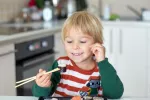 Niño sujeta un trozo de sushi con palillos Un niño sonriente se dispone a comer un plato de sushi en la cocina de su casa
