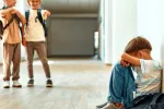 Niño sentado en el suelo de la escuela acosado por dos compañeros Niño sentado en el suelo se tapa la cara mientras dos compañeros de escuela se ríen de él
