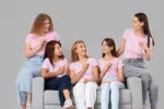 Cinco mujeres jóvenes con camisetas y cintas rosa que simbolizan la lucha contra el cáncer de mama Grupo de mujeres jóvenes con cintas rosadas contra el cáncer de mama sentadas en un sofá