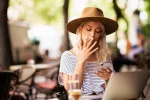 Prohibición de fumar en terrazas exteriores Mujer joven rubia con sombrero fuma un cigarrillo en una terraza y escribe mensajes de texto