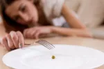 Chica joven con malnutrición muy delgada Joven muy delgada a punto de pinchar un guisante de un plato