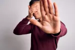 Joven muestra su rechazo a saber y se cubre los ojos con una mano Joven se tapa los ojos con una mano mientras con la otra hace un gesto de rechazo