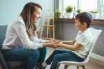 Terapia escolar para prevenir el TDAH en niños Mujer psicóloga coge de las manos a un niño sentado en una silla que sonríe