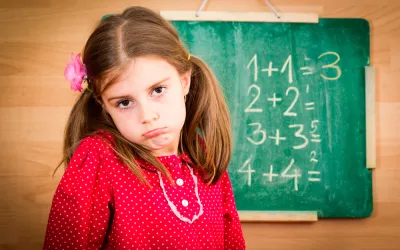 Niña con gesto compungido junto a una pizarra con operaciones matemáticas